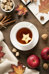 A cup of fruit tea, a light warm blanket, yellow and orange leaves, apples, spices on a brown wooden table. Hugge, autumn cozy mood. Top view.