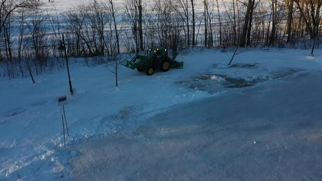 Aerial drone view coming down showing a landscape of frozen river, trees and a snow removal tractor removing snow near a skating ring at Berges-des-quatres-vents in Laval-Ouest