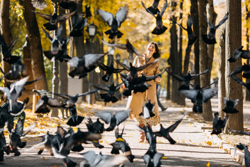 Happy young woman feeding pigeons in a city central park on warm autumn day.