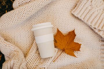Reusable cup of coffee on a white knitted sweater, with an orange autumn leaf.