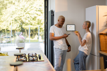 Multiracial couple controlling smart home devices with a digital tablet on the wall. Concept of...