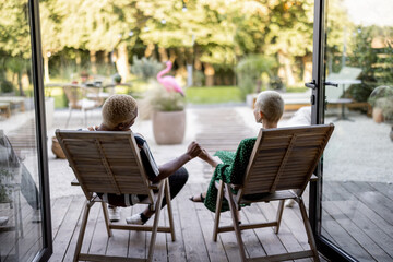 Multiracial couple drinking cocktails while sitting on wooden chairs at home terrace. European girl and black man spending time together. Concept of leisure. Modern domestic lifestyle