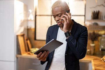 Latin man using digital tablet while talk on smartphone. Concept of modern domestic lifestyle. Young focused guy work remotely from home. Interior of kitchen in modern apartment