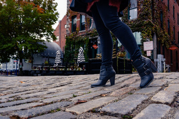 A young women walk down a cobblestone street in jeans and black boots.