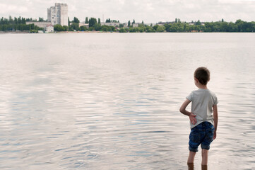 A child with brown hair and a light T-shirt and rolled-up jeans is standing with his back to his knees in the river with a view of the city with a place for text