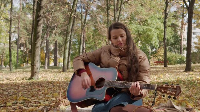Young romantic talented beautiful girl woman composer musician with long hair plays guitar sings song sitting On Red Plaid Yellow Fallen Leaves Look At Camera Cold Weather In Fall Park. Autumn Concept