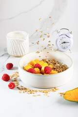 A bowl of granola topped with fresh mango cubes and raspberries, served on a white marble surface with milk.