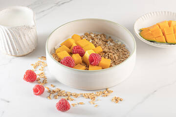 A bowl of granola topped with fresh mango cubes and raspberries, served on a white marble surface with milk.