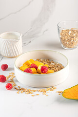 Bowl of granola with yogurt, milk and fresh raspberries, blueberries, mango for a healthy breakfast on a white marble table