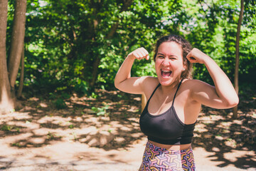 Woman in sportswear making strength gesture and smiling. Young woman in gym outfit outdoors making strength and victory gesture smiling. Photo with space for text.
