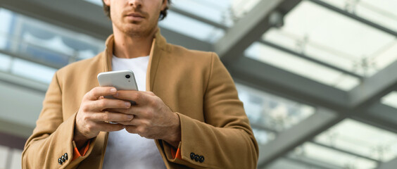 Cropped view of smartphone in hands of blurred man in coat outdoors, banner  © Dmytro Hai
