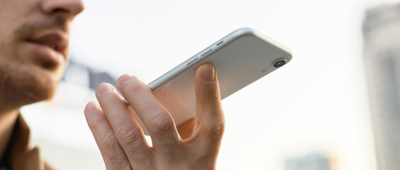 Cropped view of smartphone in hand of man recording voice message outdoors in evening, panoramic shot 