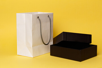 The concept of Valentine's Day. Black Friday. An open stylish black gift box on a yellow background lies next to a white paper bag. Packaging for expensive gifts. Close-up. Front view