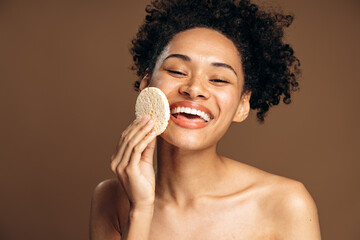 Portrait of smiling woman keeping sponge in her arm while closed her eyes against brown background. Cosmetology, beauty and spa concept