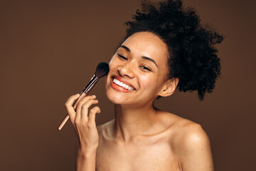 Close up of brunette lady holding make up brush near her face and smiling toothy to the camera. Beauty procedures concept. Stock photo
