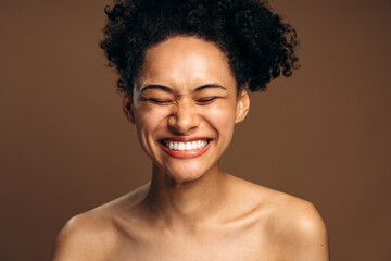 Brunette woman with curly hair laughing out loud with closed eyes while posing at the studio over...