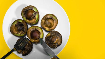 Bad Avocado halfs placed on plate with fork and knife Yellow background. Rotten Food Contaminated Avocado Necomestibile Fruits Wasting Food Unhealthy Tropical Fruits. Food concept Copy space.