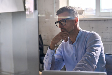 Serious focused European broker trader in glasses looking at pc laptop working at desk in office. Focused male ceo manager leader analysing financial data using computer. Shot through glass.