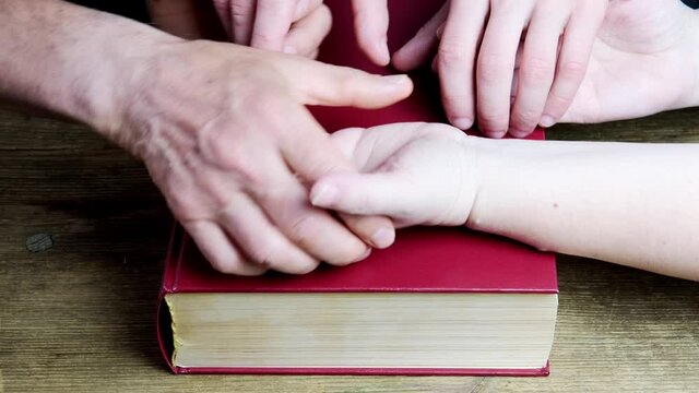 close-up of the hands of two men and a woman folded in a prayer gesture on a thick family bible in a dark red cover, the concept of eternal Christian values, the unity of people in faith