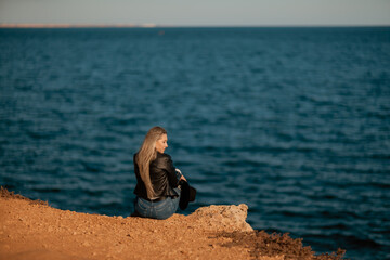A blonde girl in a stylish black leather jacket is sitting with her back to the seashore.