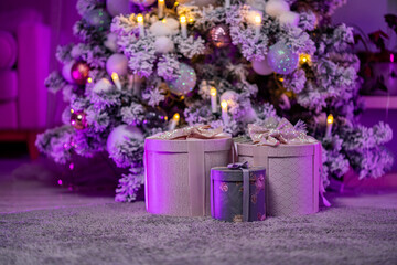 Boxes with holiday gifts for the New Year are decorated with balls, snowballs, sparkling candles of a snow-covered tree. Christmas morning in the interior with bright purple backlighting