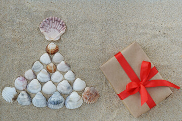 Top view of christmas tree made from sea shells and wrapped giftbox with red ribbon on light sand background< copyspace. Creativity, christmas