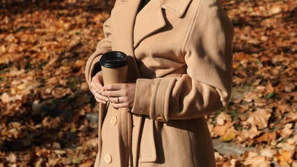 Smiling young curly hair woman in trendy beige coat with cup of coffee outdoor. Beautiful young woman drinks coffee in the autumn park with falling foliage.