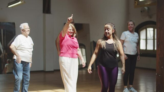 Long shot of senior people having dance class with teacher. Tracking shot of female instructor learning moves with men and women attending lesson, stepping side to side, raising arms up. Hobby concept