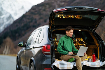A woman in a sweater is sitting in the trunk of a car and holding a laptop.