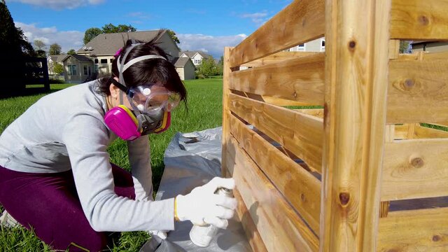 A young caucasian woman wearing face mask safety goggles and gloves is spray staining  a wooden fence outdoors. Concept video for diy project and health and safety measures she wears gas mask.