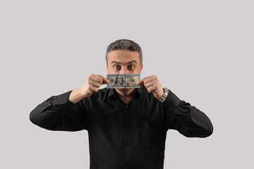 A middle-aged man in a black shirt holds a 100 American dollar bill and covers his mouth with it