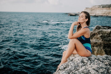 Fototapeta na wymiar A young beautiful tanned woman with long hair in blue bikini sitting at rocks near blue green clear ocean sea water. Woman relax on sunset at sea, enjoy holidays and weekend vacation in summer time.