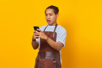 Portrait of handsome Barista who is shocked and surprised to see discount promo on smartphone on yellow background