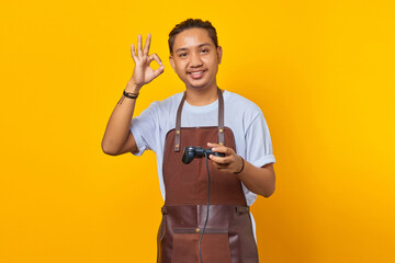 Portrait of cheerful handsome Asian young man wearing apron holding game controller and confident showing ok gesture over yellow background