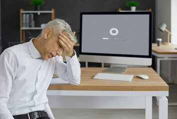Unhappy, sad senior man sitting at office desk with computer with gear icon and progress bar on monitor, holding head in despair, waiting for software update to install or new operating system to load