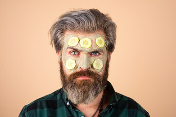 Man with clay mask and cucumber slices on face. Spa, dermatology, wellness and facial treatment concept. Man having cosmetic moisturizing mask, isolated studio background. Male face care.