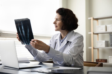 Concentrated mature female surgeon traumatologist in white uniform analyzing x-ray images, examining fracture, considering problem solution, sitting at modern workplace in clinic, medicare concept.