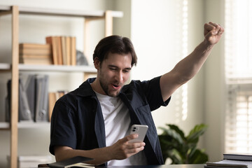 Happy young man looking at smartphone screen, celebrating online lottery gambling auction win,...