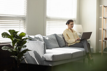 Full length happy middle aged woman using computer software application, resting on comfortable sofa, communicating online in social networks or web surfing information, modern technology addiction.