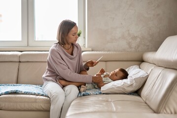 Worried young mother sitting on sofa beside her sick son with high fever. Mom measures temperature...