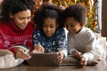 Smiling African American mom and two small children relax on floor watch funny video on tablet gadget together. Happy biracial mother and little kids have fun using modern pad on winter holidays.