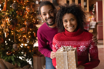 Fototapeta na wymiar Portrait of happy young African American couple pose with holiday presents near beautiful decorated Christmas tree at home. Smiling biracial man and woman exchange gifts celebrate New Year together.