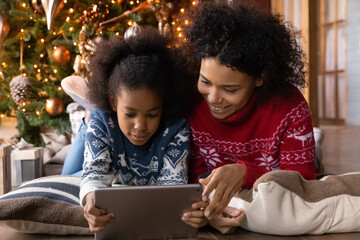 Happy biracial young mother and small daughter enjoy Christmas winter holidays using tablet gadget together. Smiling African American mom and little girl child relax at home playing on pad device.