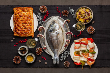 Christmas eve dinner concept. Baked dorado fish, fish pie, potato salad, seafood skewers. White wine glases. Festive decoration. Catholic tradition related to the Feast. Top view.