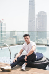 Handsome young Asian man sitting on the rooftop near the swimming pool. Chinese guy, millennials life, lifestyle in the big city, Modern Asia