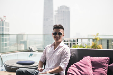 Handsome young Asian man sitting on the rooftop near the swimming pool. Chinese guy, millennials life, lifestyle in the big city, Modern Asia
