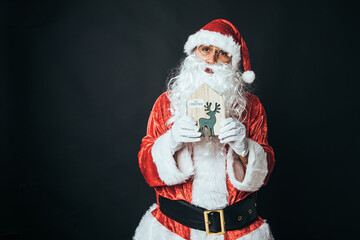 Man dressed as Santa Claus holding the figure of a green wooden reindeer, on black background. Concept of Christmas, Santa Claus, gifts, celebration.