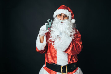 Man dressed as Santa Claus holding the figure of a green wooden reindeer, on black background. Concept of Christmas, Santa Claus, gifts, celebration.