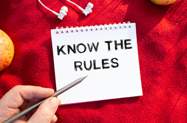 Male hand writes with a pencil in a white notebook on a red plaid in nature. Text in a notebook KNOW THE RULES.