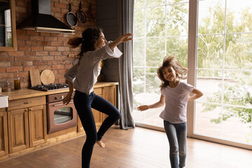 Excited crazy young mom and daughter girl dancing to music in kitchen, having fun at home together,...